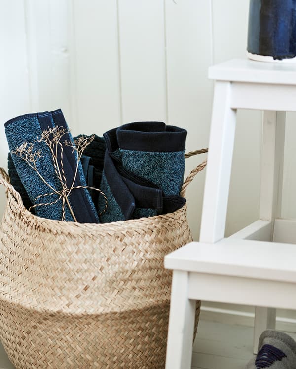 Basket made from natural woven material, placed beside a white step stool and filled with rolled up blue towels.