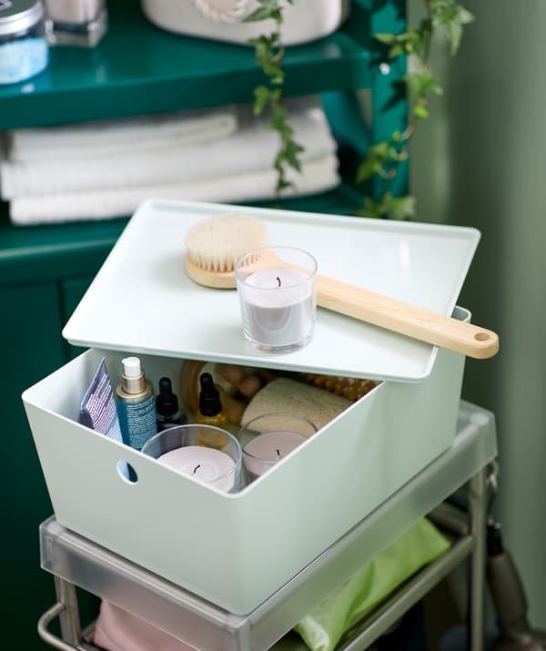 Bathroom setting with a trolley with accessories, on top a KUGGIS box holding a wellness kit: oils, brushes, SINNLIG candles.