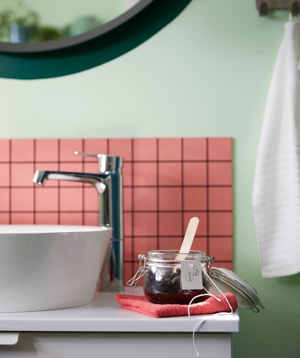 Bathroom washstand with a folded VÅGSJÖN cloth on which stands a KORKEN jar with handwritten tag, filled with homemade scrub.