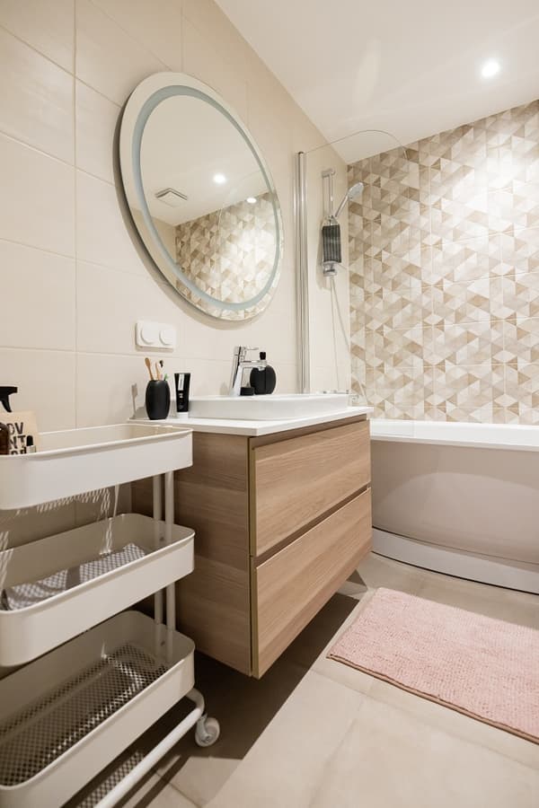 Bathroom with round mirror, wooden vanity, geometric tile wall, and pink bath mat.