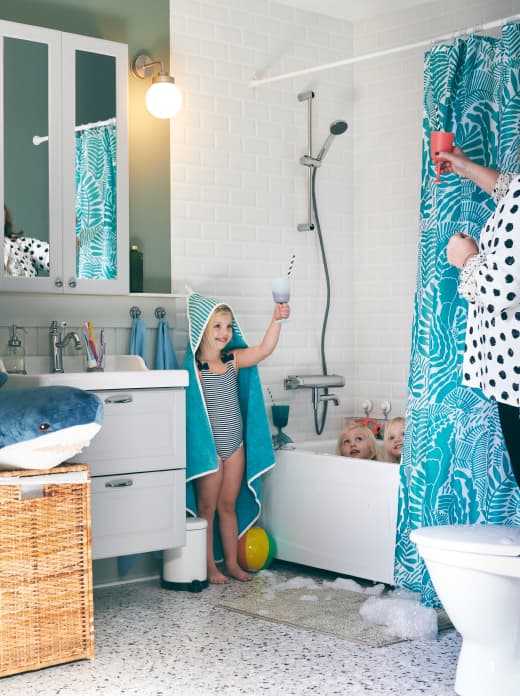Bathroom with three girls around a bath. One of them, draped in a RÖRANDE hooded towel, makes a play toast with her mother.