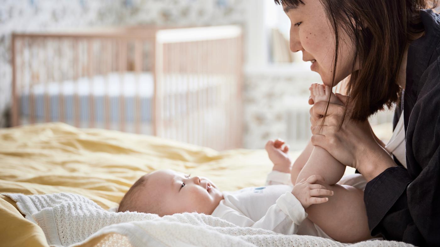 Bayi yang gembira sedang baring di atas selimut GULSPARV di atas katil dengan ibunya memegang kakinya dan tersenyum padanya.