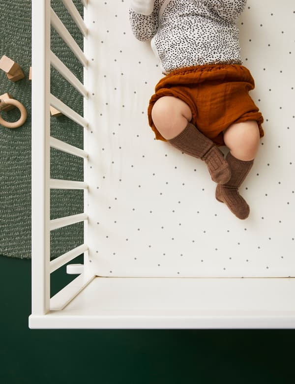 Bébé portant de jolis vêtements allongé dans un lit à barreaux dont le matelas est couvert d’un drap-housse LENAST. Il y a des jouets en bois sur le tapis, sur le sol, à proximité.
