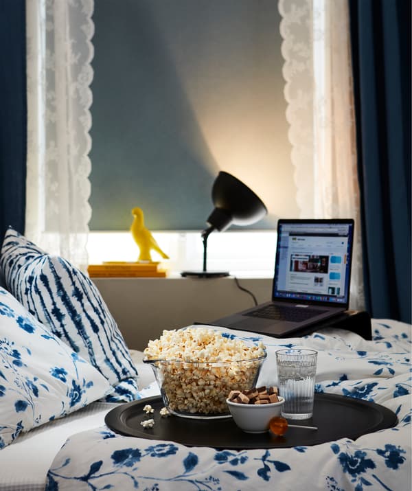 Bedroom turned simple film theatre; laptop on a stand, lowered black-out blind, lit lamp facing away, snack tray on the bed.
