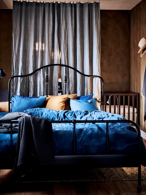 Bedroom with a black SAGSTUA bed and, in a corner behind a room-dividing curtain, stands a wooden baby cot.