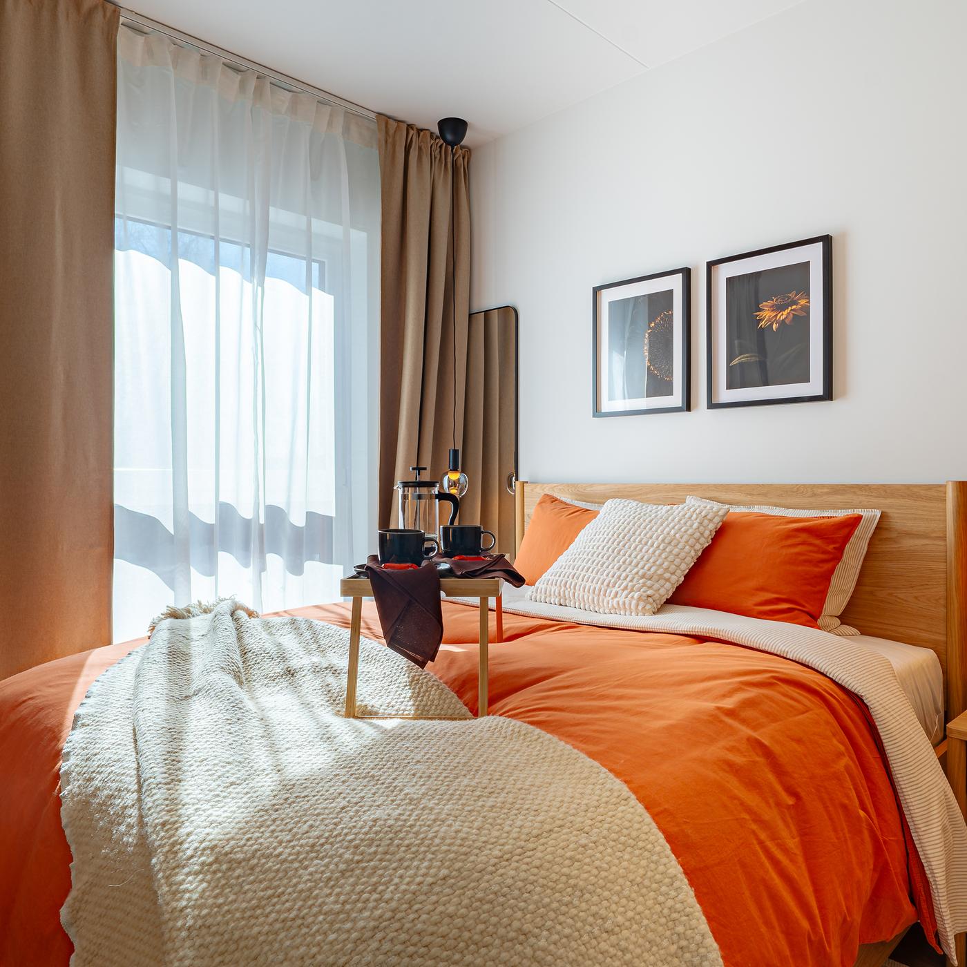 Bedroom with an oak veneer bed frame and bedside tables, dressed in terracotta and neutral textiles, featuring mirrors on each side of the bed, soft curtains, and pendant lamps above the bedside tables.