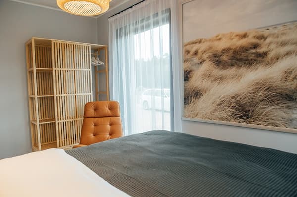 Bedroom with natural wood wardrobe and leather accent chair – Light-filled bedroom with a wooden open wardrobe, tan leather chair, and large wall art of coastal sand dunes.