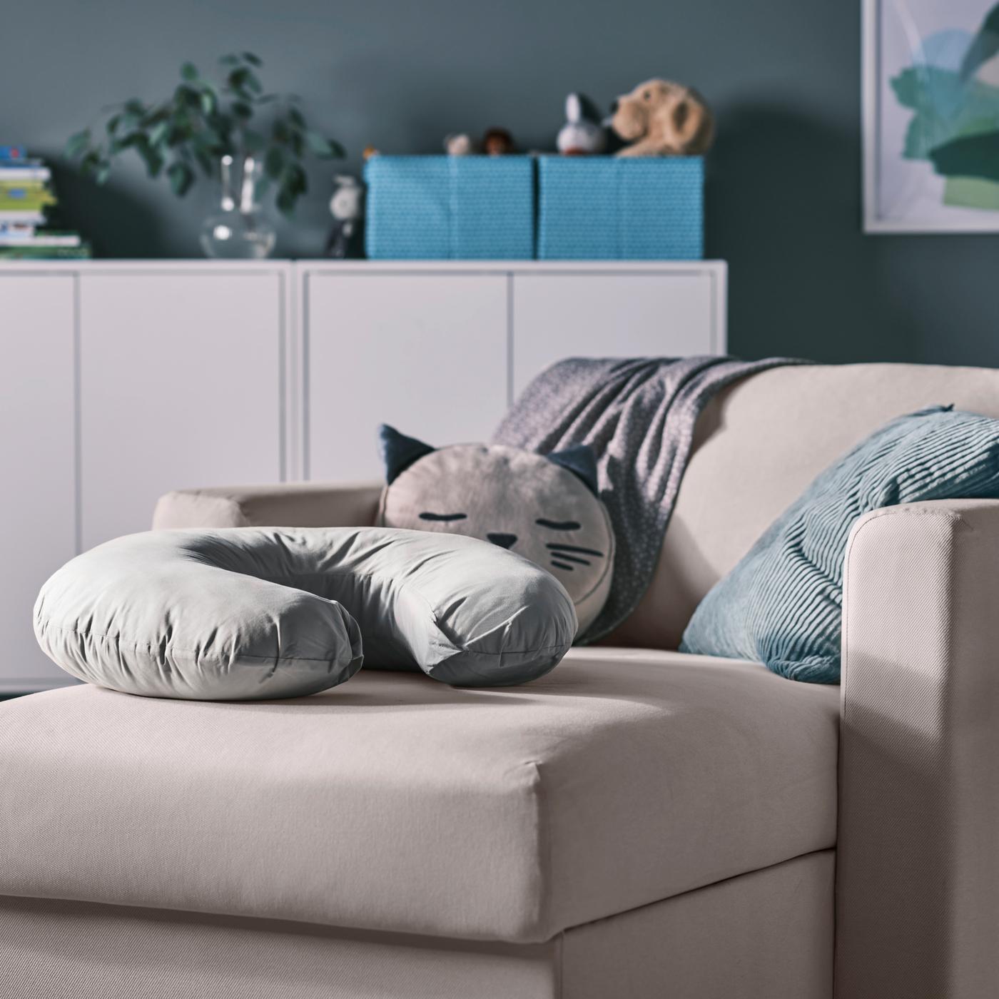 Beige chaise longue met grijs verzorgingskussen en diverse kussens, plus witte kasten met dozen en een vaas bovenop.