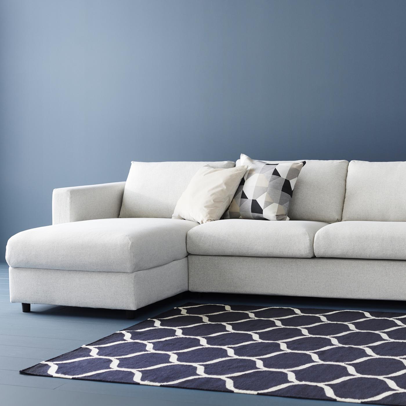 Beige VIMLE sofa bed with chaise longue against a blue wall, with blue patterned rug. There are two cushions on the sofa.