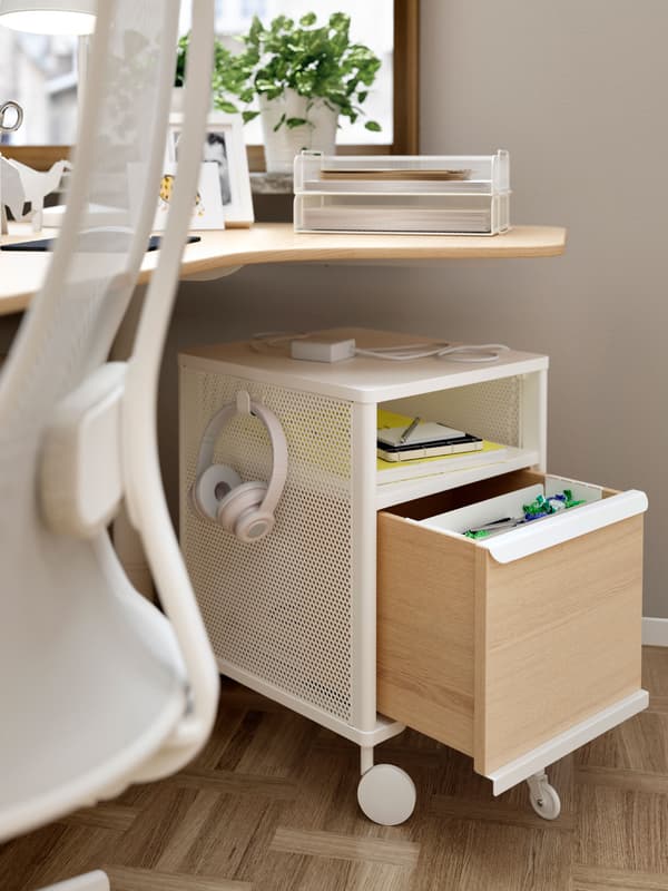 BEKANT storage unit under a desk with an open drawer and objects on the shelf. Headphones hang from a hook on the side.