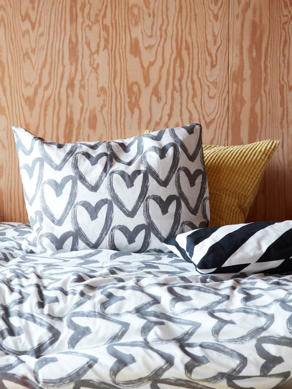 Beli i sivi LYKTFIBBLA jorgan sa šarom u obliku srca na krevetu u spavaćoj sobi s drvenim panelima.