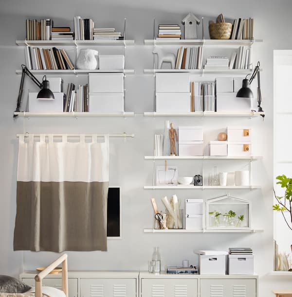 Beli zid napunjen policama, TJENA kutije za odlaganje i IKEA PS ormarići, svi u beloj nijansi i postavljeni simetrično.