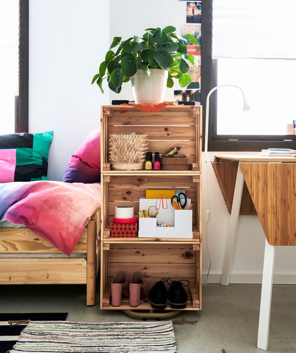 Bibliothèque peu remplie constituée de trois caisses en bois empilées entre un lit et une petite table.