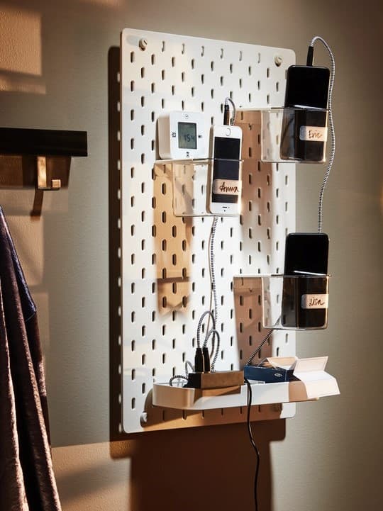 Bílá dírkovaná nástěnka IKEA SKÅDIS na stěně s průhlednými úložnými boxy, ve kterých se nabíjejí mobilní telefony pomocí kabelů.