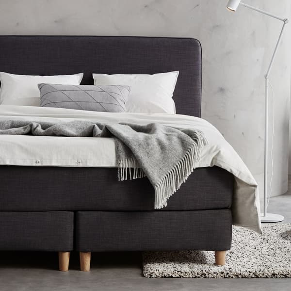 Bilik tidur berwarna kelabu dengan katil dipan DUNVIK berwarna kelabu gelap dilitupi linen katil berwarna putih. Lampu lantai/bacaan NYMÅNE berwarna putih di sebelahnya.