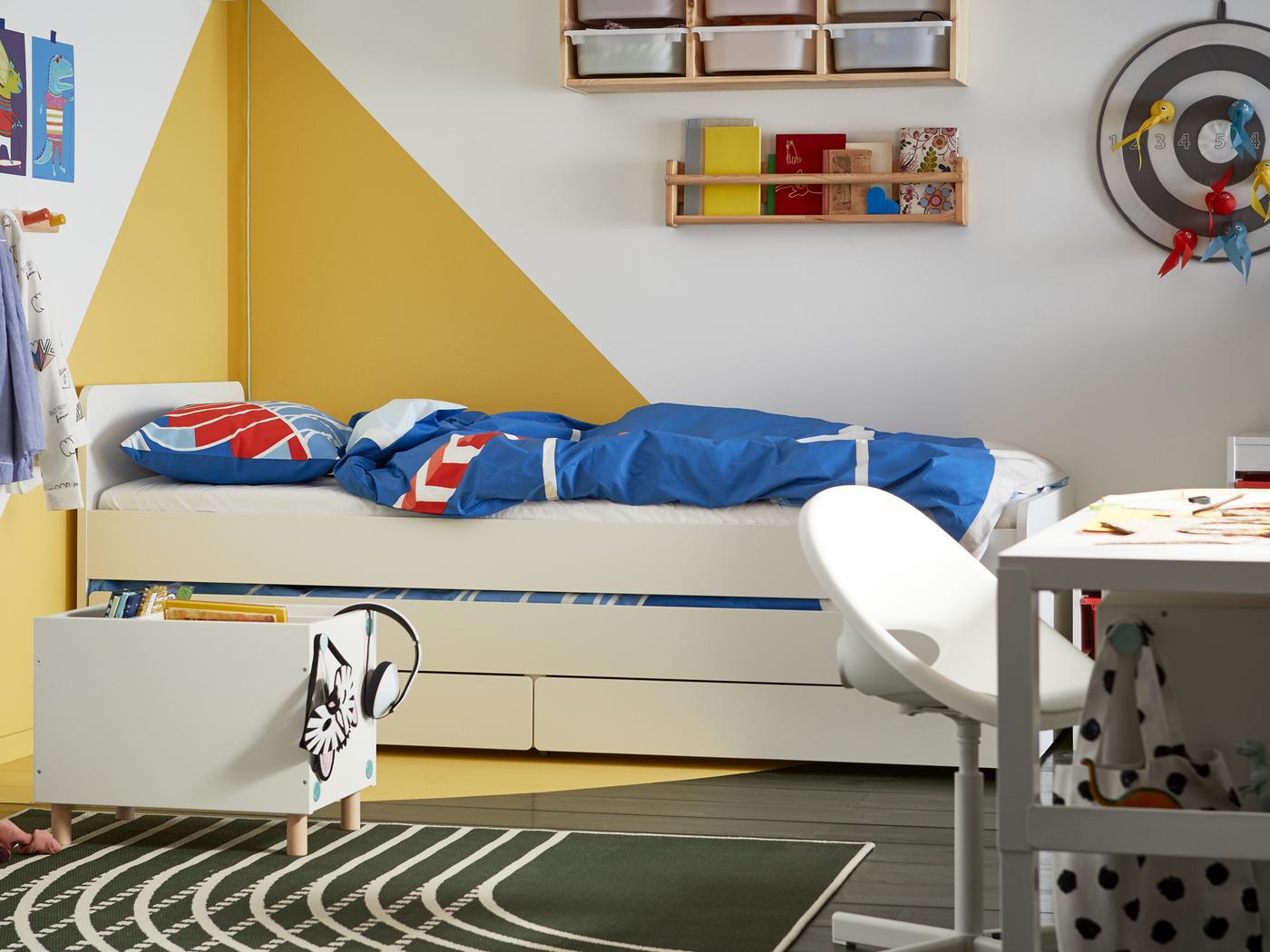 Bilik tidur kanak-kanak dengan dinding grafik berwarna putih dan kuning, katil berwarna putih dengan storan di bawahnya, linen katil berwarna biru dan merah.