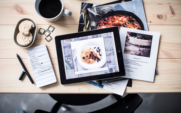 Bird’s-eye view of cooking inspiration on a tablet and in magazines, set at a table with coffee, note pad and pen.