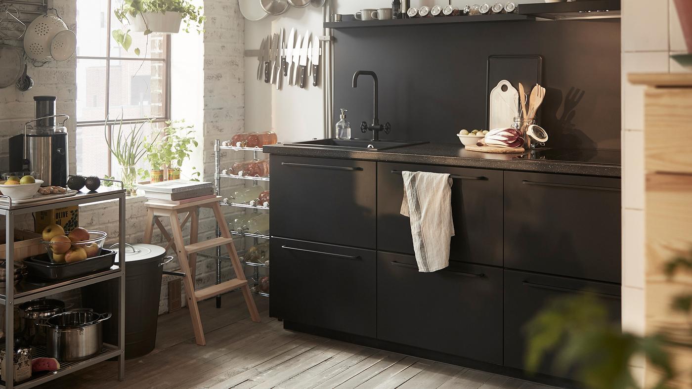 Black kitchen cupboards with a sink, a metal trolley and utensils stored on rails by a window.