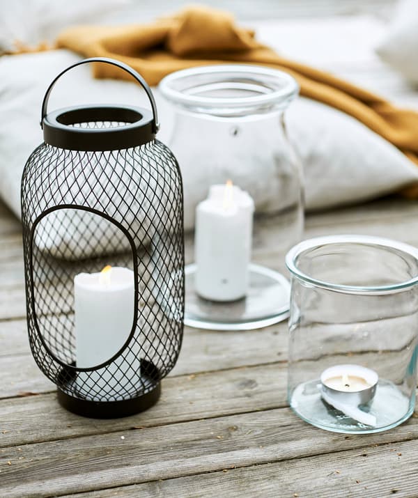 Black mesh wire lantern with block candle beside two glass jars with candles, next to a cushion and yellow throw on decking.