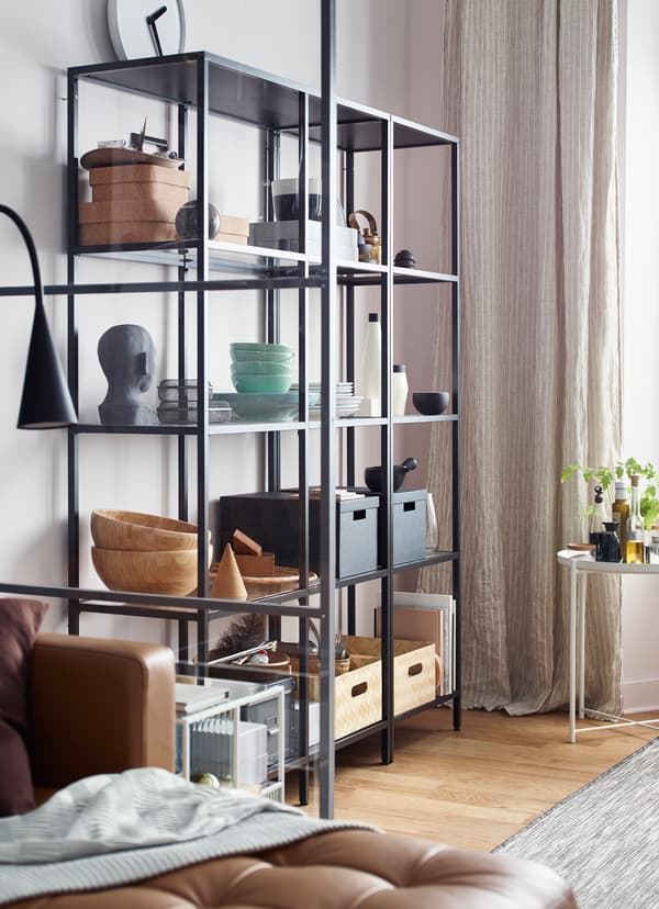 Black wired open storage shelf, holding boxes, bowls and statues in a dining room area.