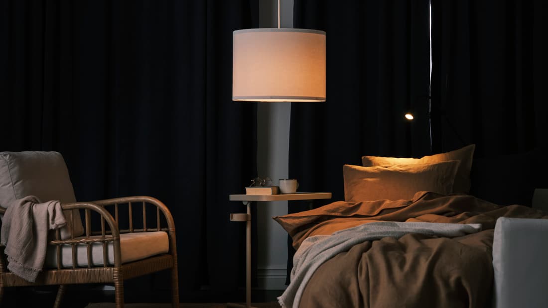 BLÅHUVA curtains block out any light from a window. A wooden chair sits a the corner of the room and a standing lamp is seen in the middle of the picture with a bed on the right.