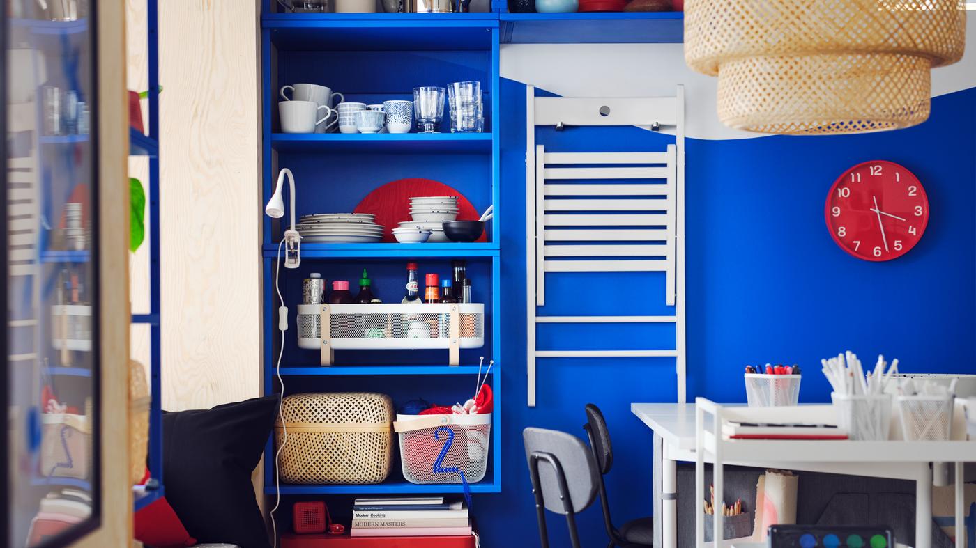 Blauwe PLATSA open stellingkast is aan een wand gemonteerd met ernaast een witte FRÖSVI klapstoel opgehangen aan haken ernaast.