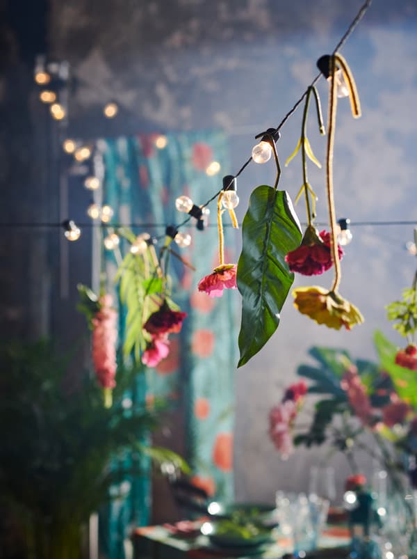 BLÖTSNÖ lighting chain with lone SMYCKA artificial flowers hanging at intervals from it, strung over a decorated set table.