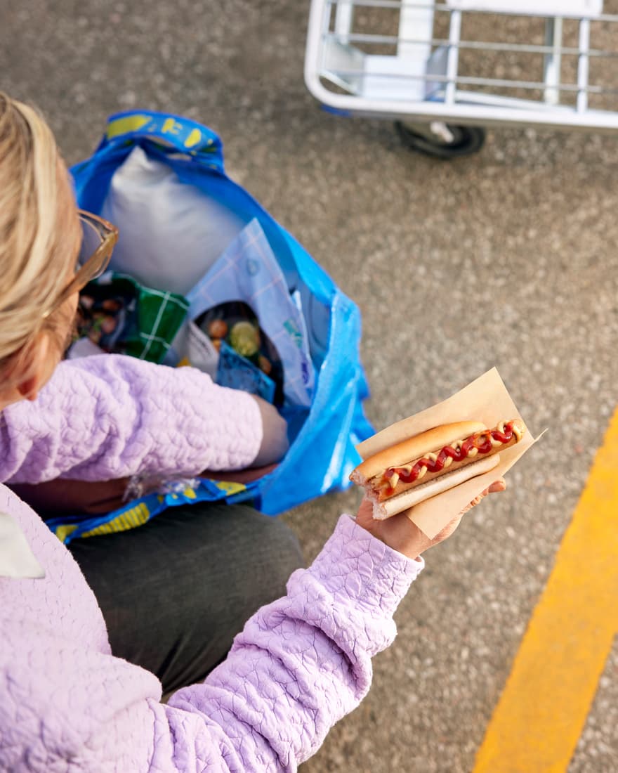 Blonde vrouw zoekt iets in een IKEA FRAKTA zak terwijl ze een IKEA hotdog met ketchup vasthoudt.