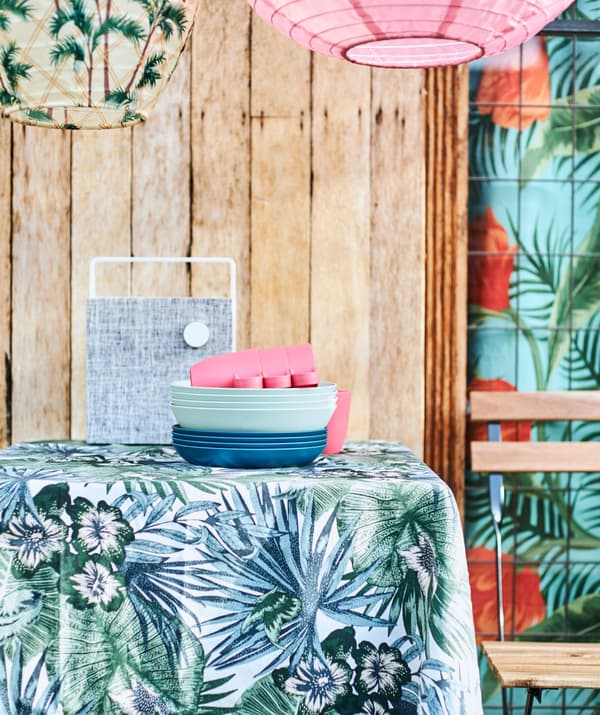 Blue and green leaf fabric covering a table, a stack of blue plates, a white speaker and a patio chair plus hanging lanterns.
