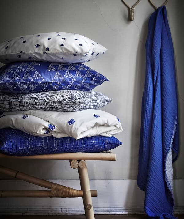 Blue and white patterned TÄNKVÄRD cushions piled up on a rattan bench and blue fabric hanging from a hook.
