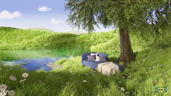 Blue BLÅKULLEN bed frame with corner headboard, a throw and cushions on top, rests in lush green grass by a pond and a tree.