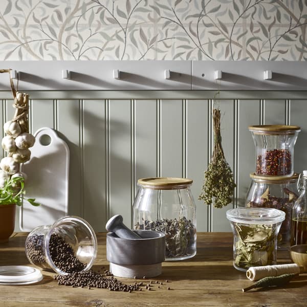 Bocaux en verre et couvercle en bois placés sur un comptoir, des herbes aromatiques sèchent à proximité.