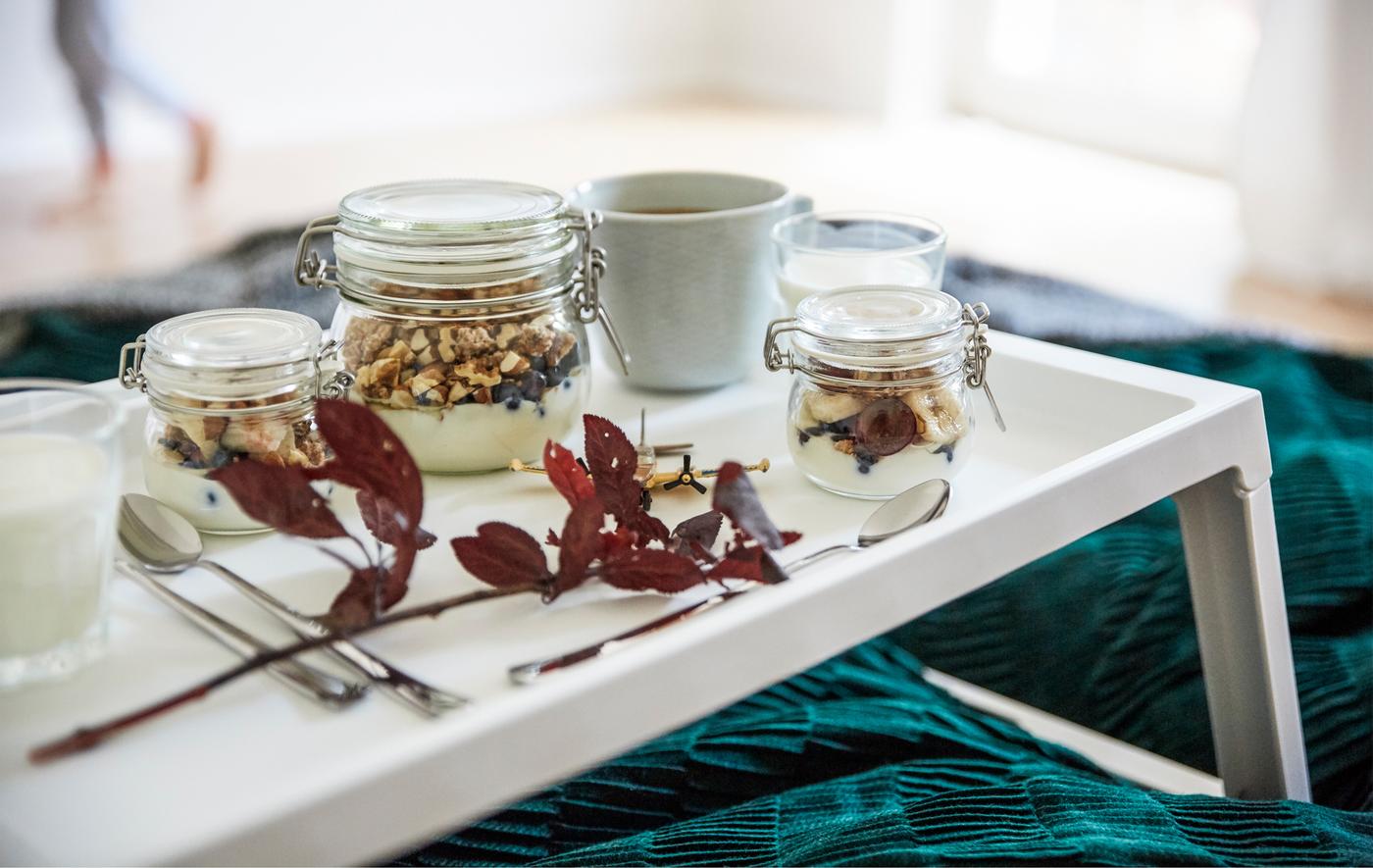 Bocaux remplis de granola et de yaourts sur un plateau blanc déposé sur un lit.