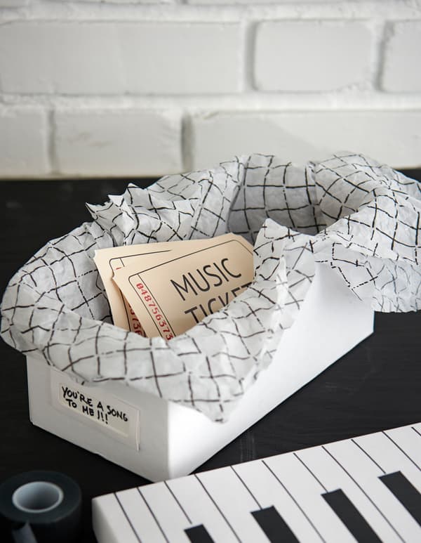 Boîte blanche décorée comme un clavier de piano, tapissée de papier de soie décoratif, contenant deux billets pour un concert. 
