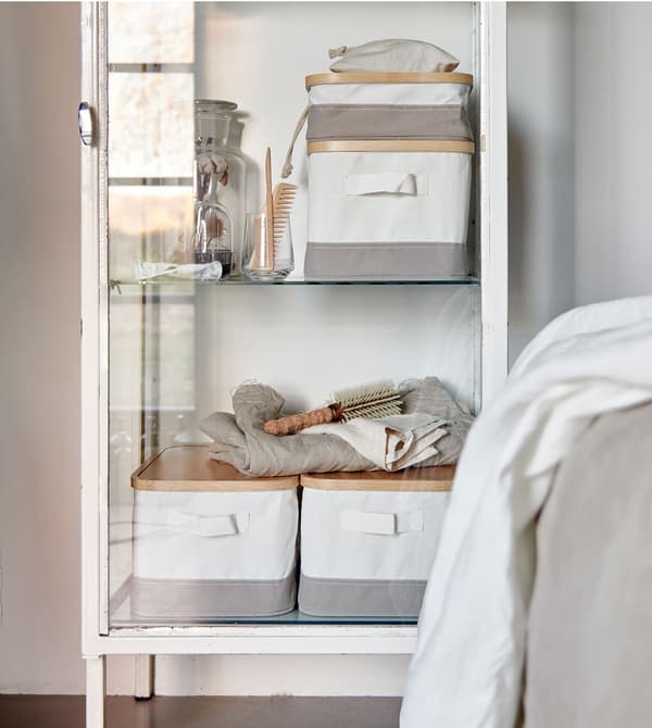 Boîtes de rangement blanches et grises RABBLA faites en bambou et en tissu, placées dans une vitrine vintage blanche.