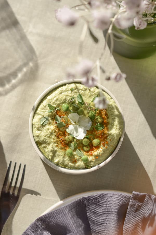 Bol de dip vert avec pois, micro-pousses et fleur sur nappe claire, fourchette et serviette au premier plan.