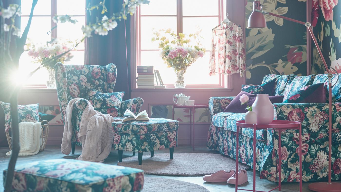Bold floral patterned sofa, armchair, footstool and cushions in a living room with a bold floral patterned wallpaper.