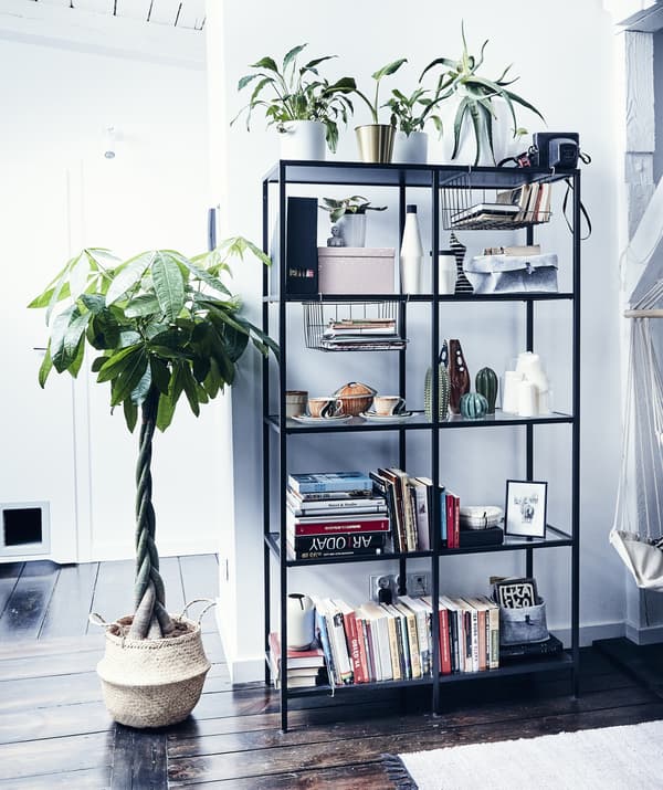 Books, plants, and ornaments on a black open storage unit next to a large plant in a rattan basket.