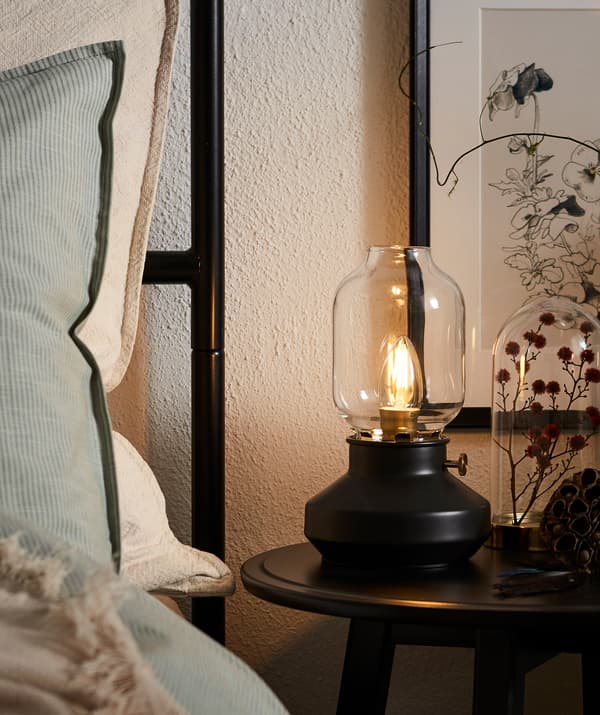 Bord d’un lit fait avec tête de lit haute, des oreillers qui y sont adossés; table de chevet ronde avec une lampe LED en forme de lampe à huile.