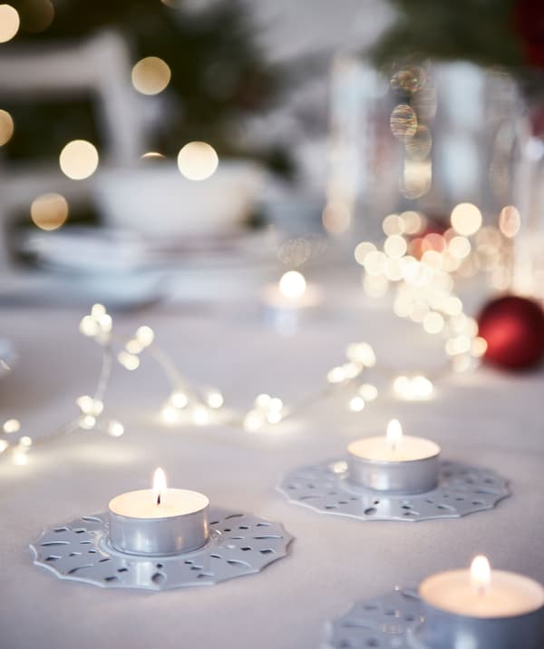Bougies chauffe-plat sur bougeoirs décoratifs placés sur la table; en flou d’arrière-plan, une grande table dressée pour le repas de Noël.