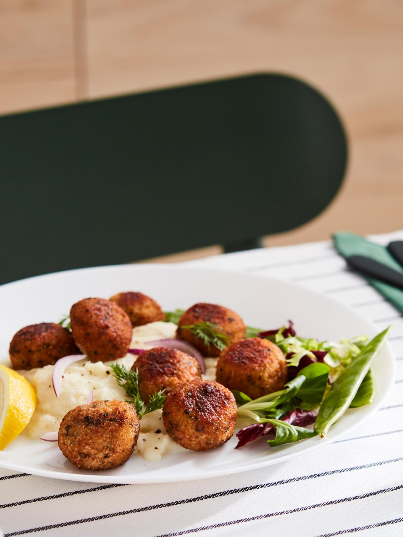 Boulettes de saumon et de cabillaud dans une assiette blanche avec de la purée de pommes de terre et de la salade, sur une table habillée d’une nappe bleue et blanche.