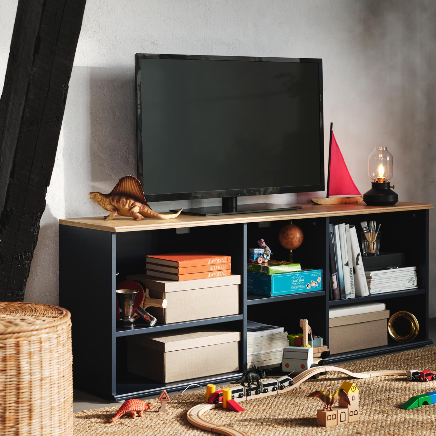 Boxes, books and toys are in the open shelves of a black-blue SKRUVBY TV bench, on which stands a black TV.