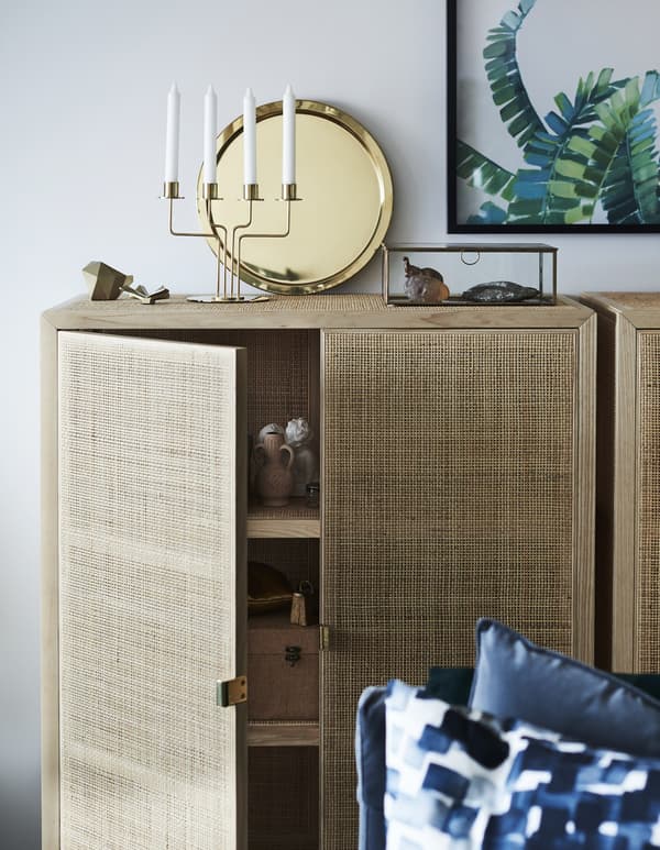 Brass-coloured tray and candle holder atop a wooden cabinet with woven fibre doors.