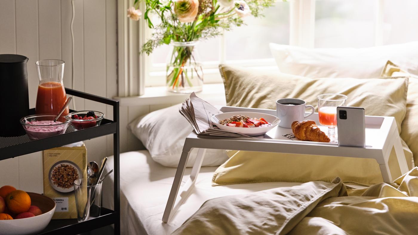 Breakfast served on a white KLIPSK bed tray on a bed made with ÄNGSLILJA bed linen, with more food on a trolley.