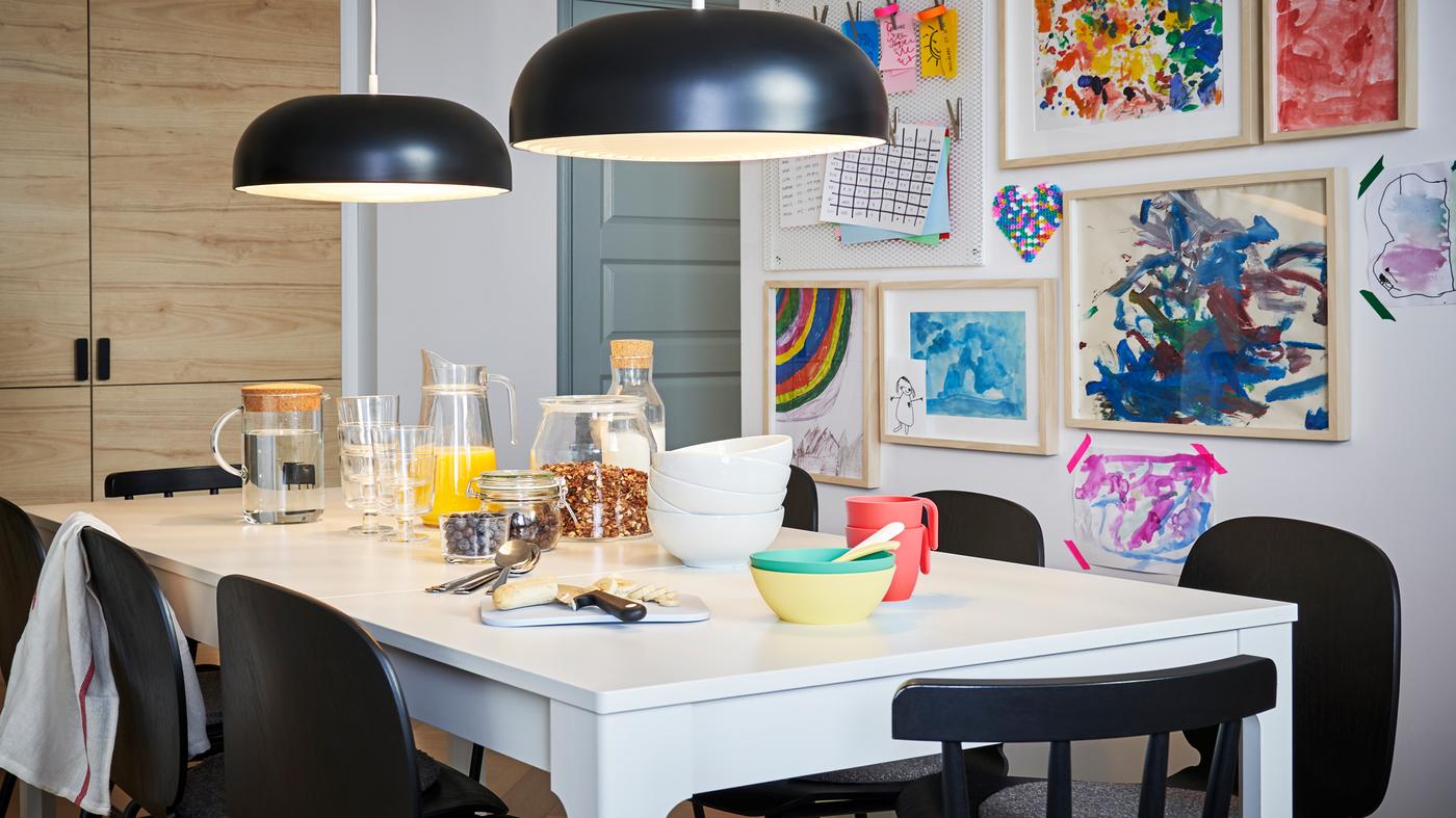 Breakfast set on a white EKEDALEN table with black chairs and two black pendants, children’s drawings on a wall behind.