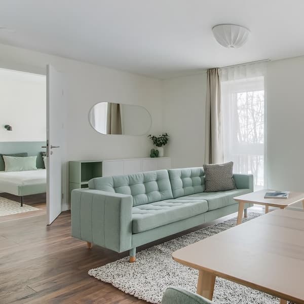 Bright and airy modern living room with a pastel green upholstered sofa centered on a textured white rug. A rectangular wooden coffee table sits in front of the sofa. On the left, a doorway opens to a bedroom with a matching green bed frame. The room features a white wall-mounted cabinet, a round-edged mirror, and a small green vase with greenery. Sheer white curtains with beige drapes allow natural light to filter through a large window, enhancing the soft and minimalist Scandinavian-style décor.