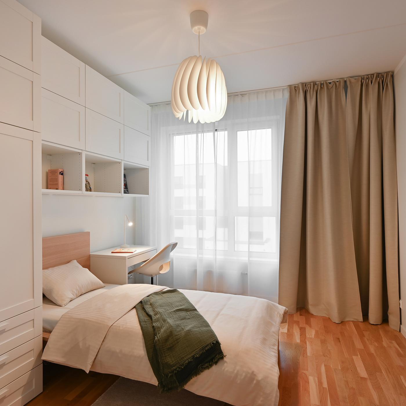 Bright and elegant bedroom with built-in white wardrobes, a compact workspace, soft neutral textiles, warm wooden flooring, and large windows bringing natural light into the contemporary space.