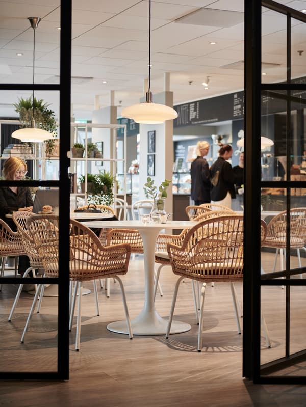 Bright cafe interior seen through glass doors, with round tables, rattan chairs, hanging lamps and a counter in the background where customers stand.