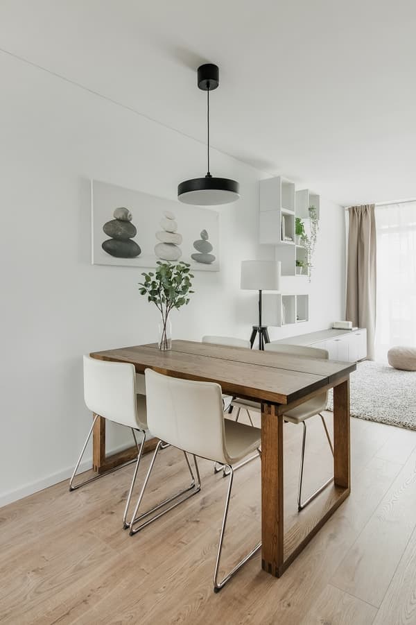 Bright dining area featuring a wooden table with four white modern chairs on metal legs. A black pendant light hangs above the table, while minimalist wall decor with stacked stones and geometric shelves add a serene, contemporary touch. Light wood flooring and natural daylight complete the cozy aesthetic.