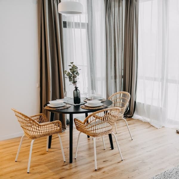 Bright dining area with round black table, wicker chairs, and floor-to-ceiling curtains.